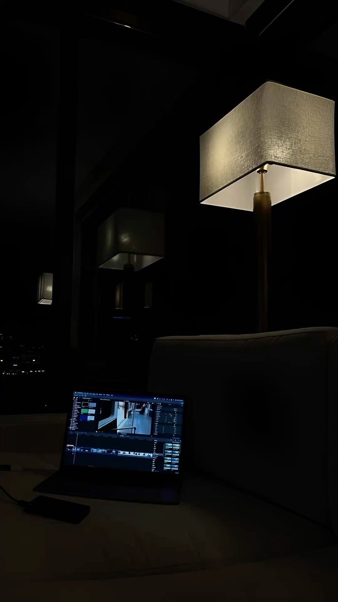 Laptop open with editing software on a sofa beside a glowing floor lamp in a dark room at night